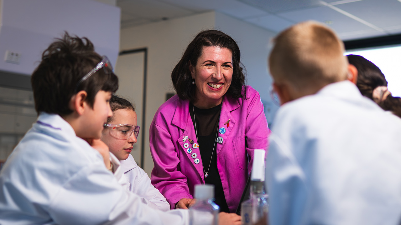 A woman in a pink lab coat smiles at students wearing goggles and white lab coats.