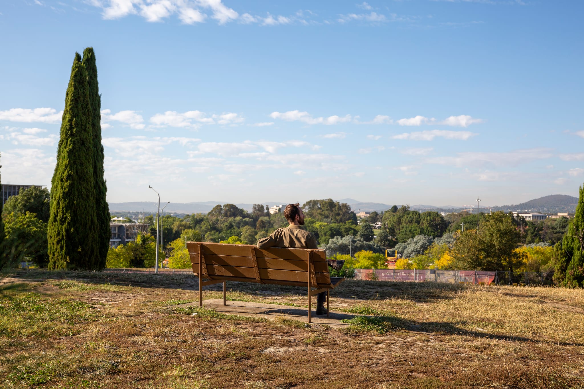 A man sitting upon a bench at the top of City Hill park overlooking Commonwealth Avenue and Lake Burley Griffen