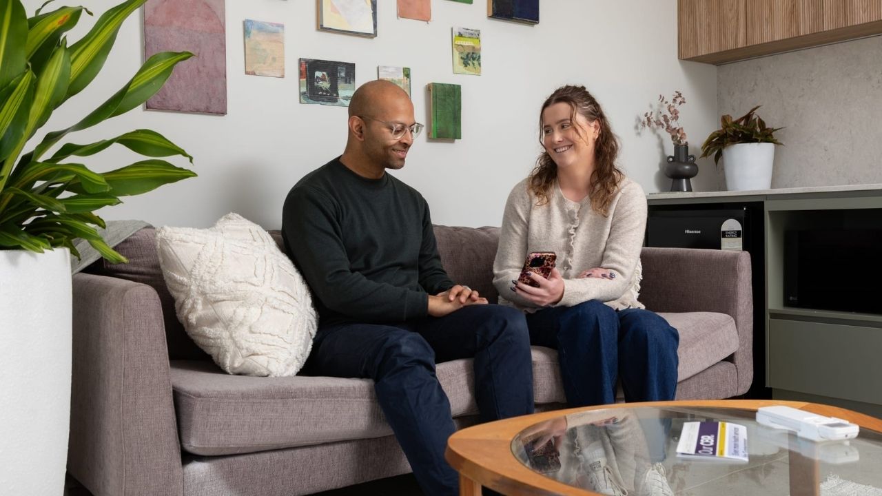 Two people sit on a light-grey sofa in a modern living room, looking at a smartphone held by one of them.
