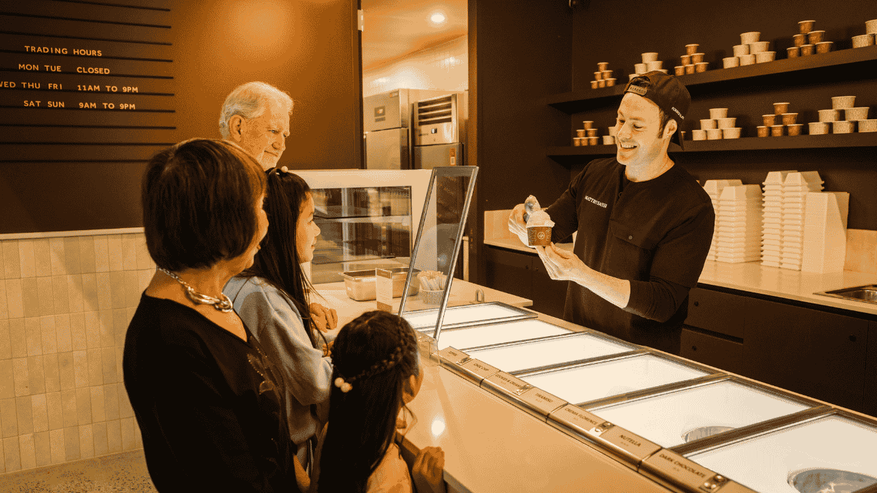 A person behind a counter scoops ice cream into a cup with people onlooking.