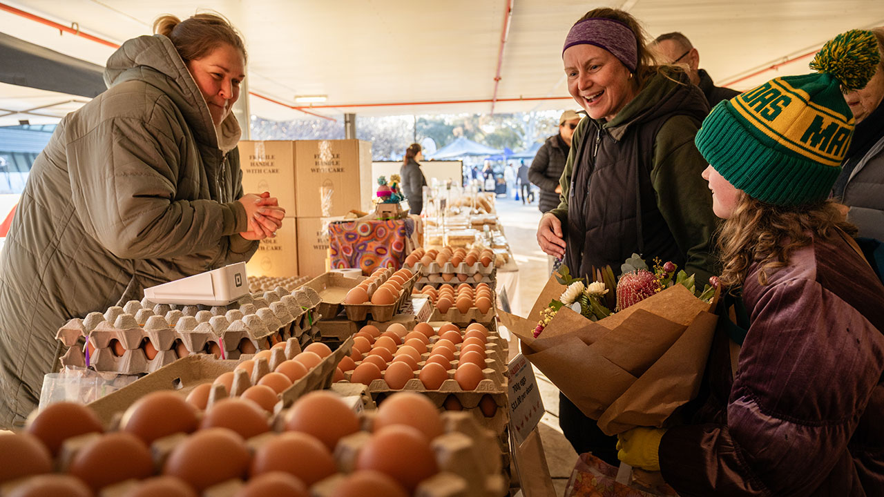 A woman stands behind a stall selling eggs at a market. Another woman and a young girl smile while looking at the produce.
