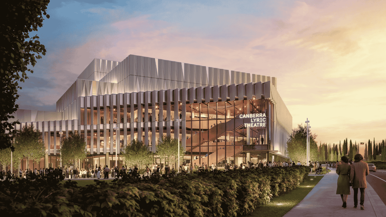 Architectural rendering of the Canberra Lyric Theatre at dusk, showing a contemporary multi-storey building with a striking silver façade of vertical fins and large glass walls. Warm interior lighting reveals open staircases and gathering spaces, while crowds of people walk and congregate around the landscaped entrance. Trees, hedges, and a broad pedestrian pathway frame the scene under a softly lit evening sky. Visible signage reads “CANBERRA LYRIC THEATRE.”
