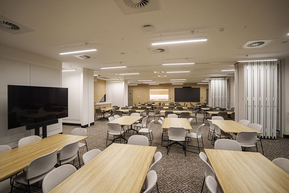 Large open multipurpose room with rows of rectangular wooden tables and light-coloured chairs, a wall-mounted screen at the front, bright ceiling lights, and movable partition walls, set up for meetings, classes or community events.
