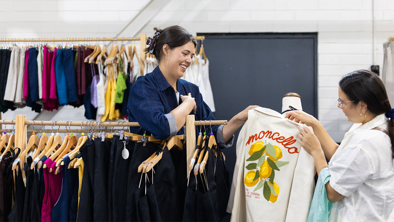 Two women hold a shirt, looking at it. One woman leans on a rack of clothing with another rack behind her.