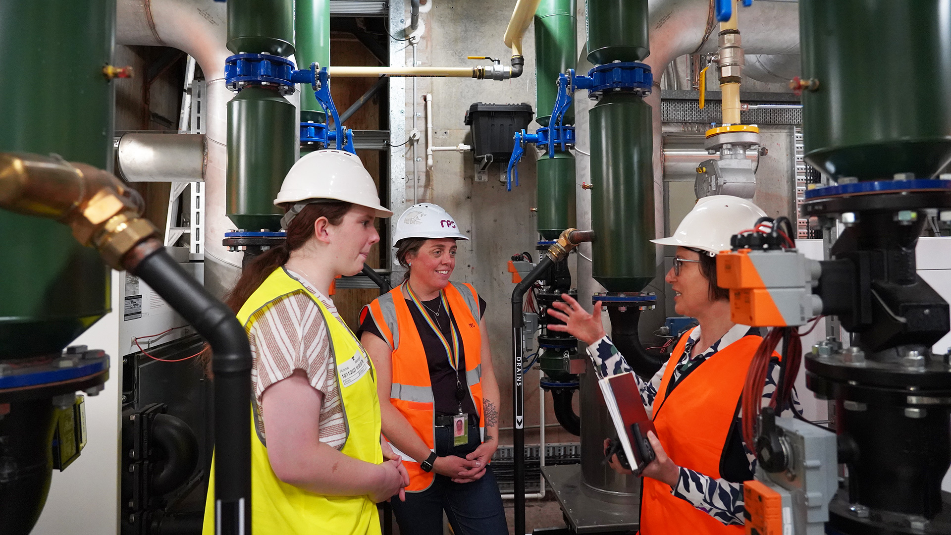 Three women in high-visibility vests and hard hats stand amongst machinery, talking.