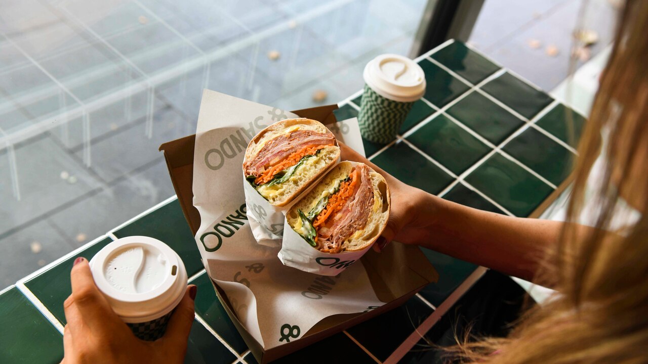 A woman holds a sandwich and a coffe at a cafe counter.