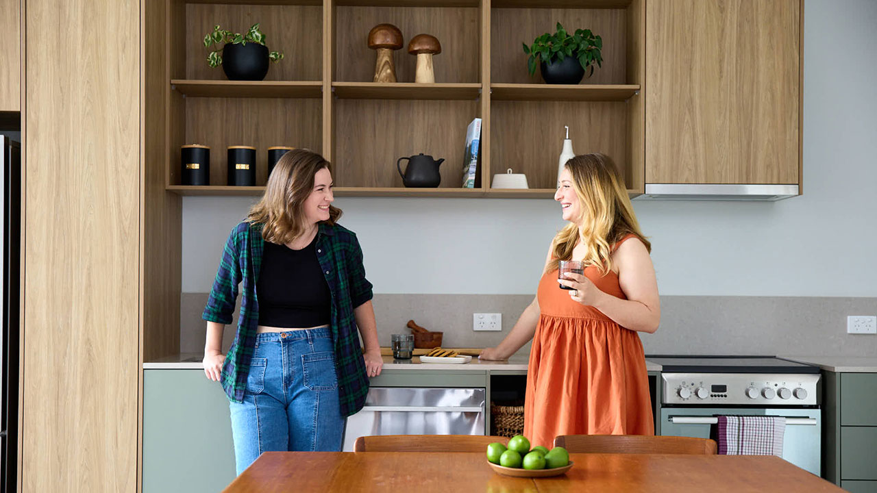 Two women stand in a modern kitchen chatting and laughing. One holds a glass of water.