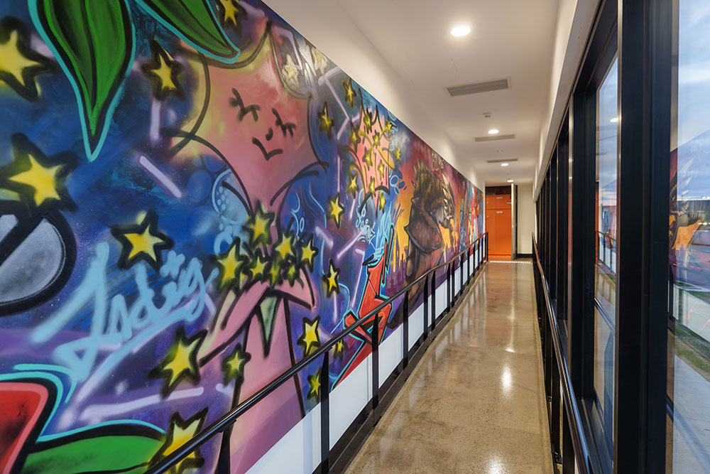 Long, narrow hallway with polished floors and a bright, colourful graffiti-style mural covering the entire left wall, featuring bold shapes, stars and abstract designs, with glass windows lining the opposite side and ceiling lights running above.