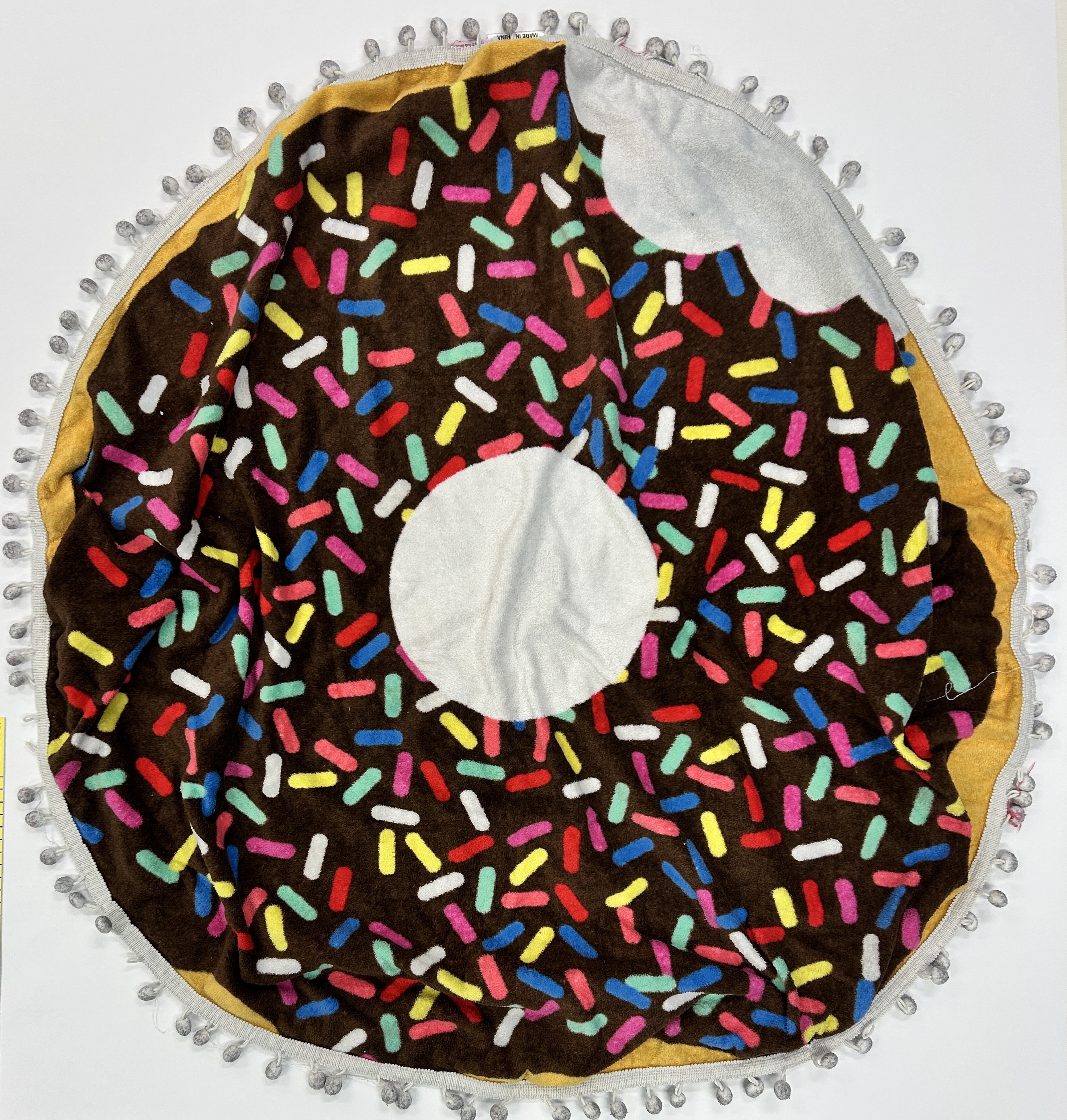 A circular towel with an image of a chocolate donut with rainbow sprinkles on it and a bite taken out of it. The towel has tassels around the edge and a white space in the centre where the donut hole is