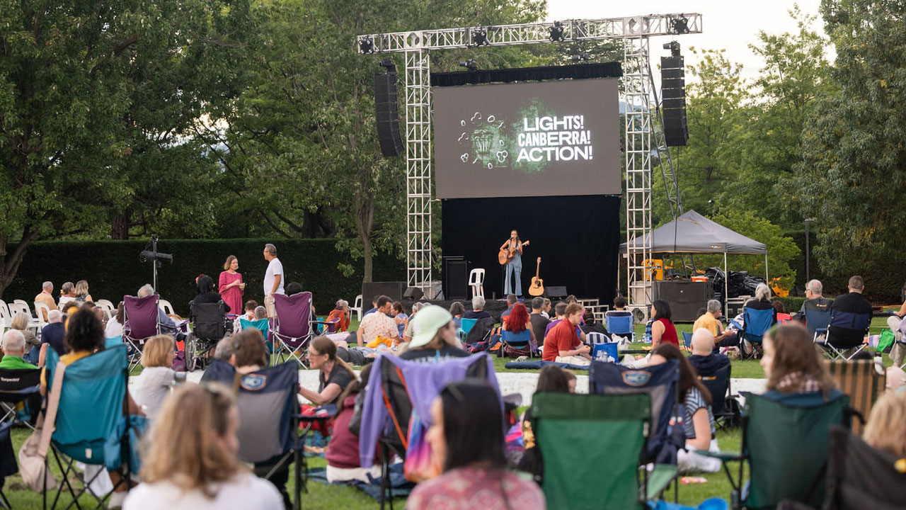 People sit on picnic rugs and folding chairs on a grassy lawn watching a live performer on an outdoor stage. A large screen above the stage displays the text “Lights! Canberra! Action!”, with trees surrounding the event space in the background.