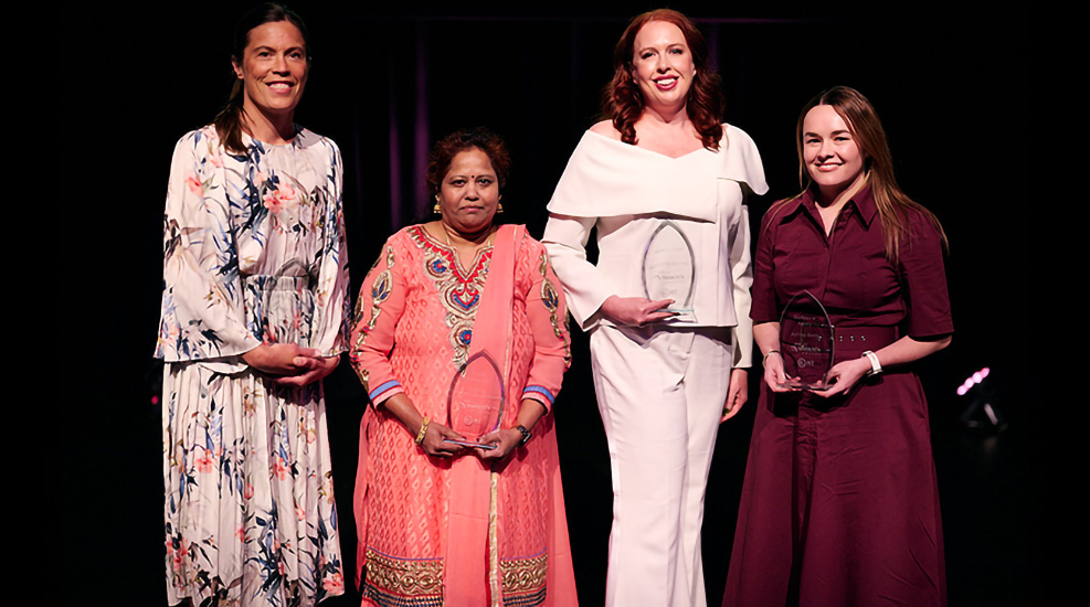 Four women standing side by side in front of a dark background. They are each holding a glass trophy.